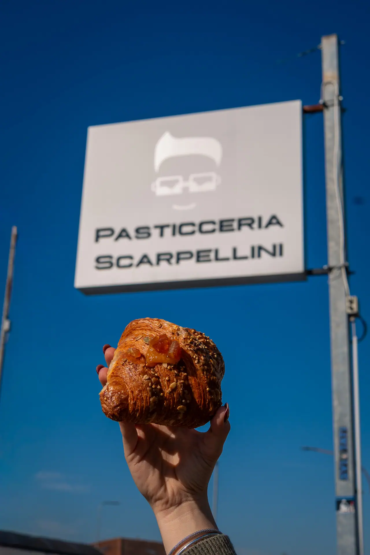 Un cornetto alzato davanti all'insegna di Pasticceria Scarpellini, cielo blu